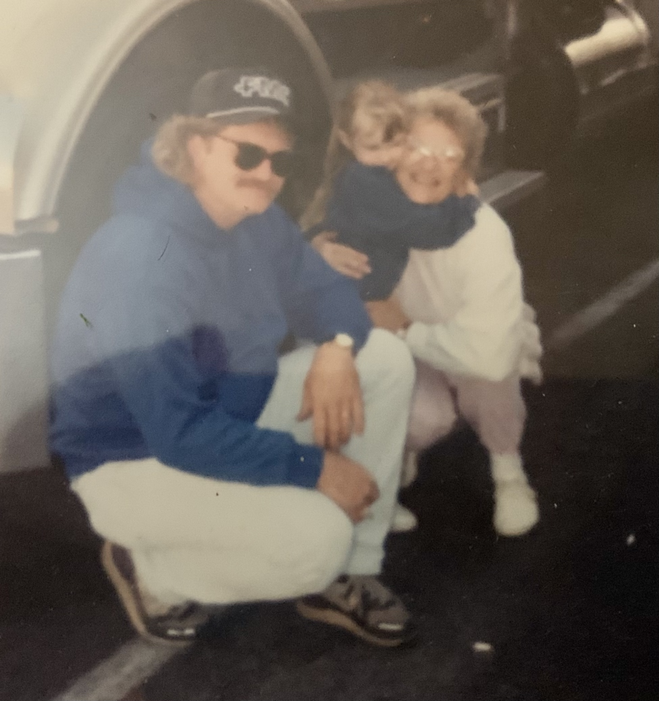 A man in a blue jacket, hat and sunglasses with curly blond hair crouches on the ground next to a young girl hugging the neck of an older woman. The girl wears blond hair in a high ponytail and a denim jacket. The older woman is mostly dressed in white with curlyl gray hair and glasses.