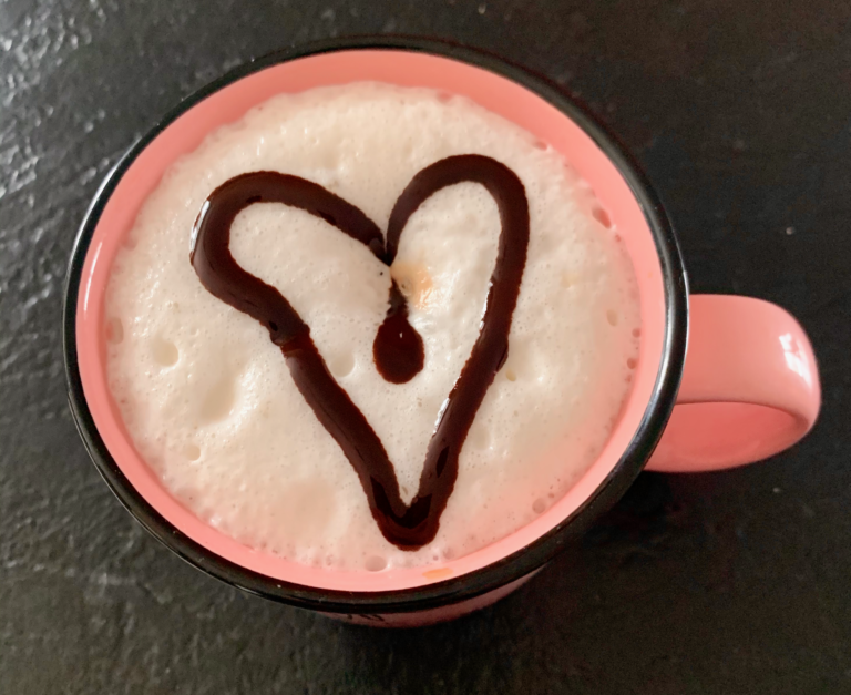 A pink coffee mug with a heart drawn in chocolate syrup on milk foam.